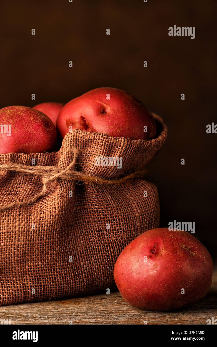 Natura morta con patate rosse. Un sacco di iuta pieno di patate rosse appena raccolte con una patata sul tavolo di legno rustico. Foto Stock