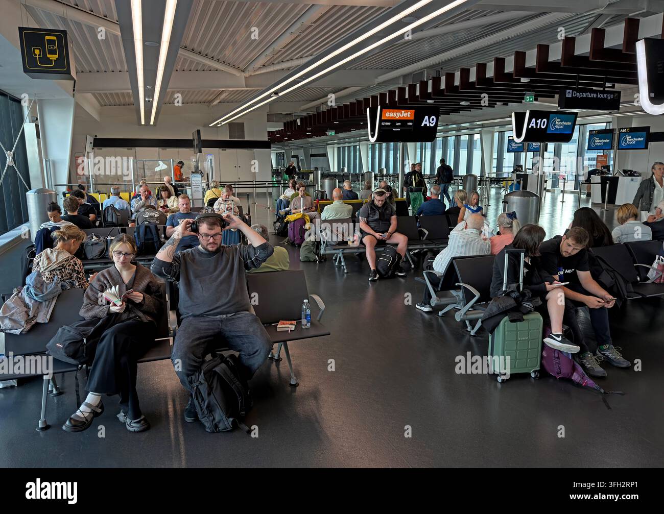 Folle di passeggeri in attesa con bagagli a mano ai gate di partenza nel Terminal 2 dell'aeroporto di Manchester durante un periodo di viaggi intenso. Foto Stock