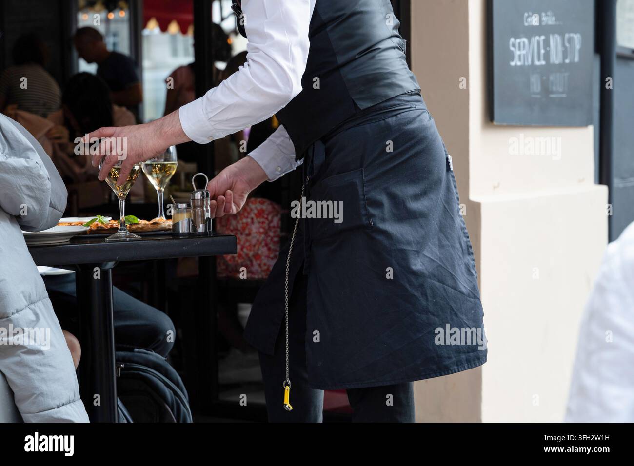 Cameriere in una brasserie parigina, che serve i tavoli sul patio, caffetteria. Piatto e bicchieri di vino bianco Foto Stock