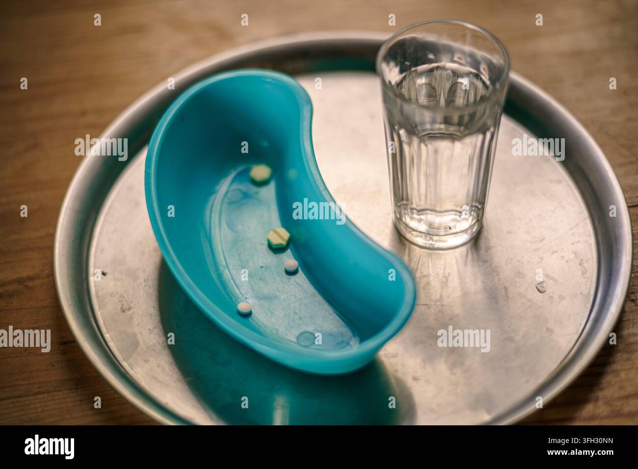 Disposizione da tavolo di varie pillole in una ciotola renale turchese accanto a un bicchiere d'acqua su un vassoio medico in metallo. Foto Stock