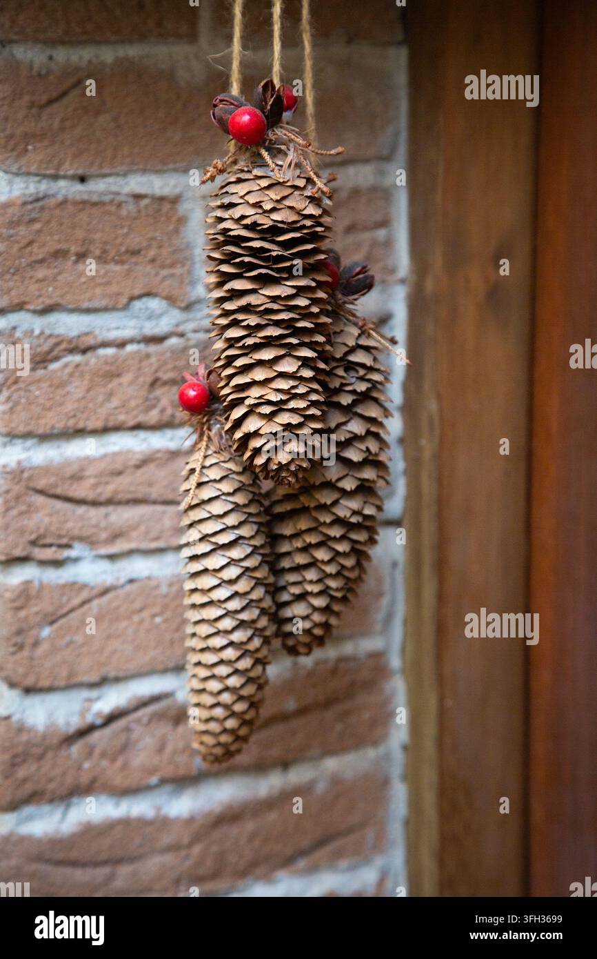 Ornamenti di pigne appesi con accenti di bacche rosse accanto a un muro di mattoni rustico e uno sfondo di legno, che mostrano una decorazione natalizia naturale artigianale con calore Foto Stock