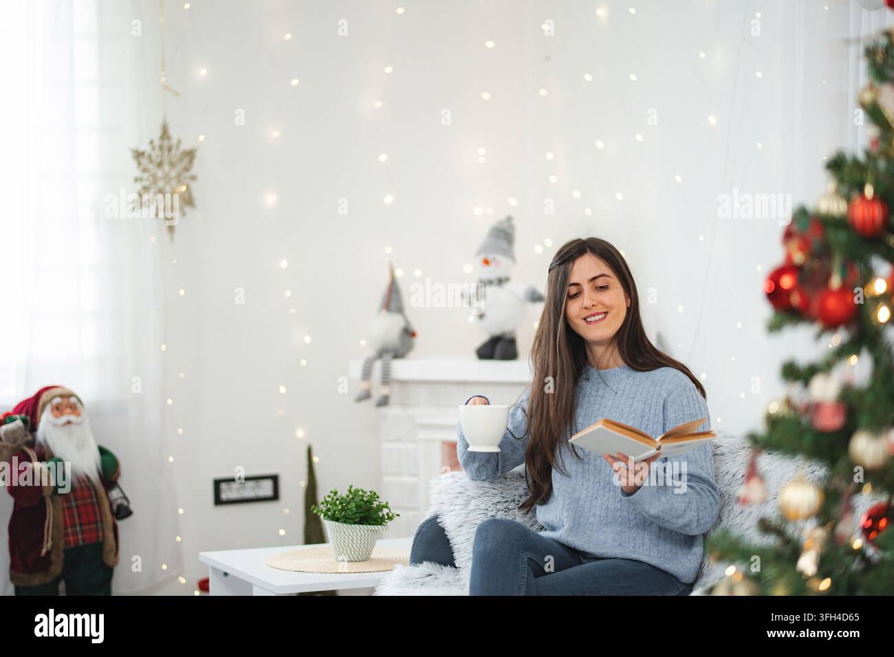 Ambientazione interna accogliente con una donna che legge un libro e si gode una bevanda calda durante la stagione delle festività Foto Stock