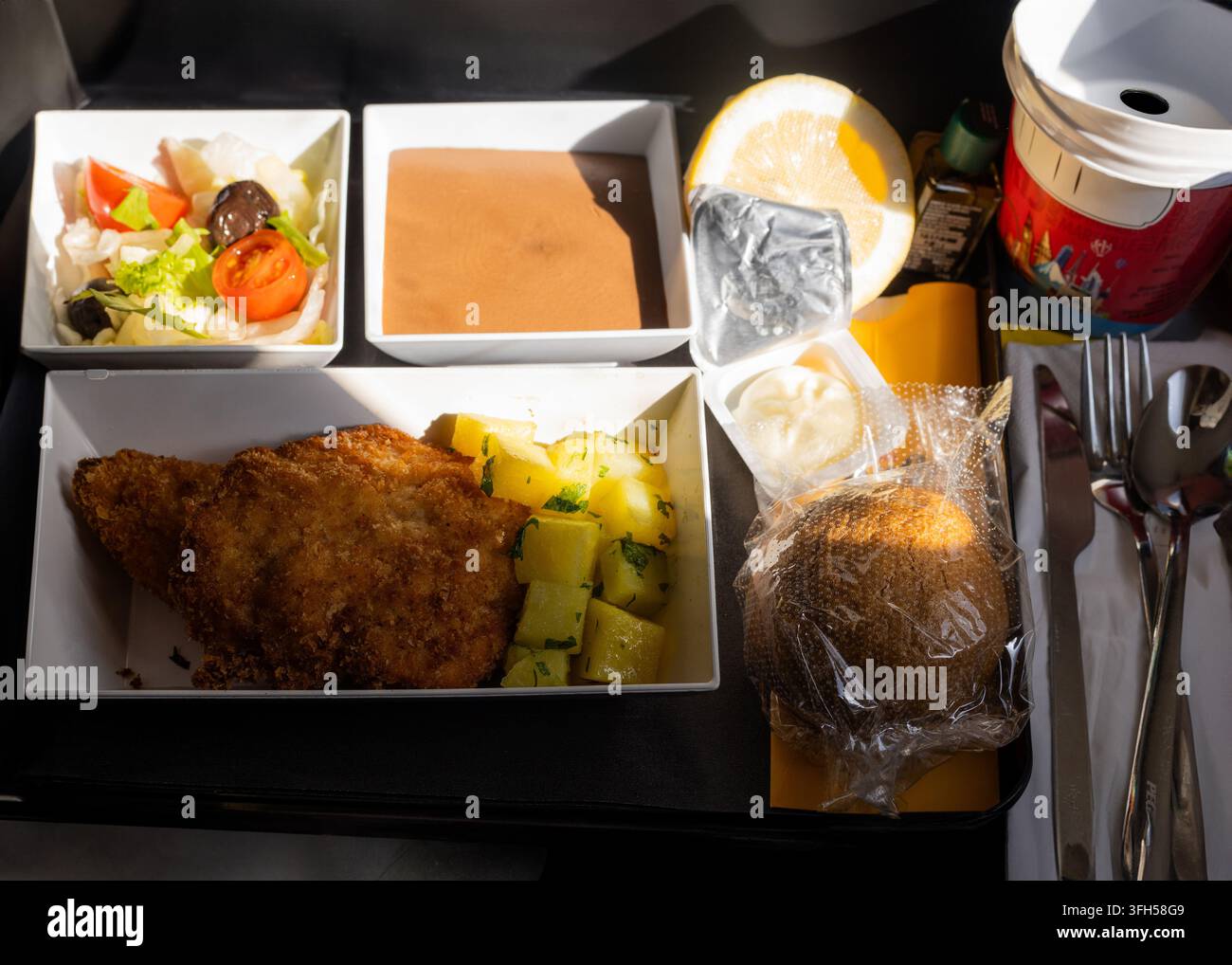 Vista ravvicinata di un pasto in aereo servito durante un volo con piatto principale caldo, patate, insalata fresca, dessert, pane, salse, limone e bevanda. Foto Stock