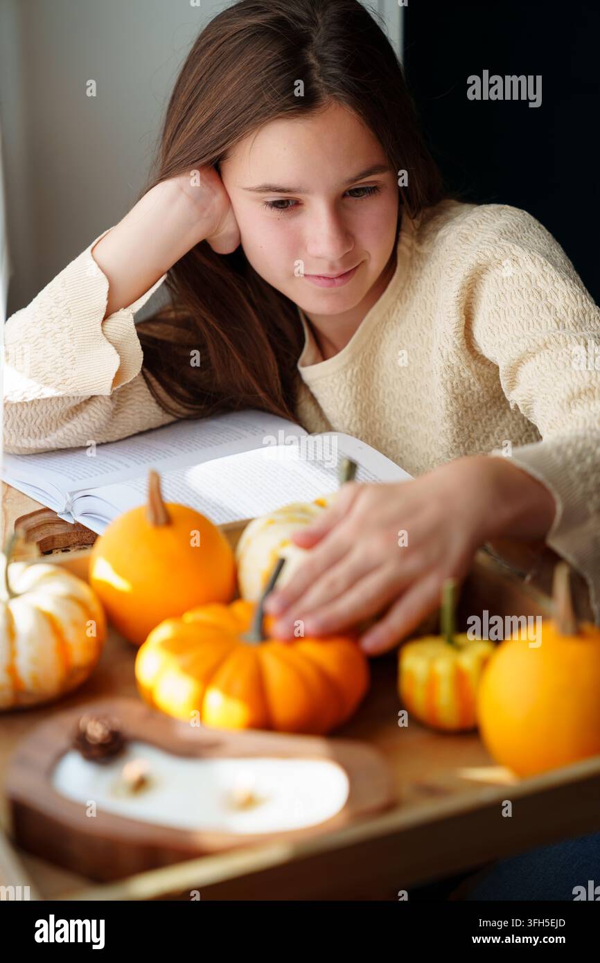 Ragazzina che tocca zucche autunnali mentre si rilassa vicino alla finestra Foto Stock