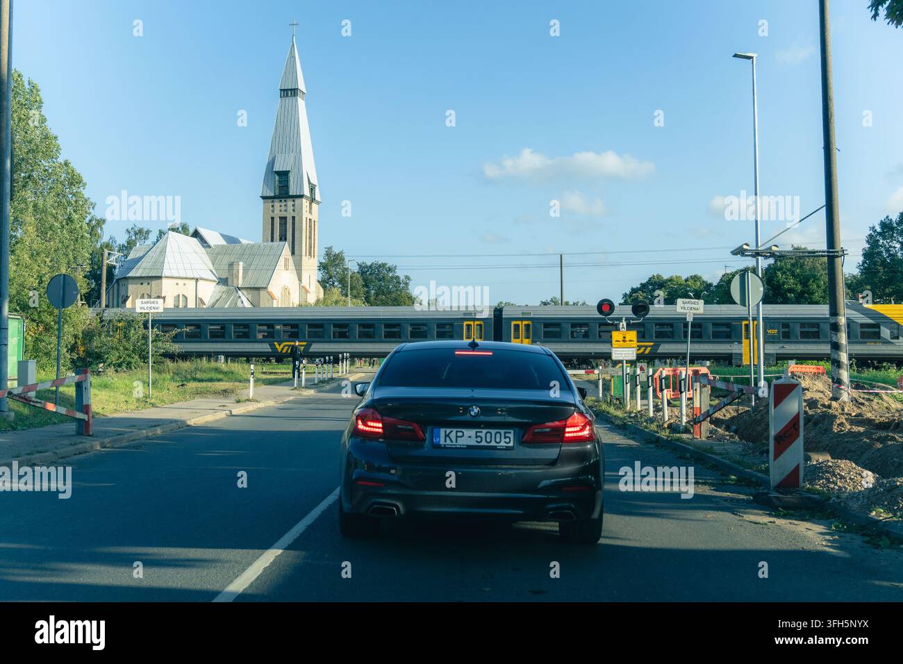 RIGA, LETTONIA - 25 AGOSTO 2026: Un treno passeggeri Vivi passa davanti a un passaggio a livello mentre le auto attendono a un segnale rosso, con una chiesa visibile sullo sfondo i Foto Stock