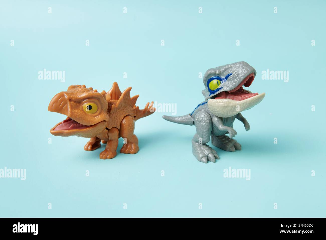 Due figure di giocattoli di dinosauri colorate posizionate fianco a fianco su uno sfondo blu pastello brillante, creando una scena preistorica giocosa e immaginativa. Foto Stock