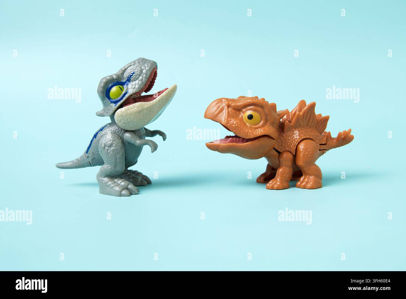 Due figure giocattolo di dinosauri colorate che si affrontano su uno sfondo blu pastello brillante, creando una scena preistorica giocosa e immaginativa. Foto Stock