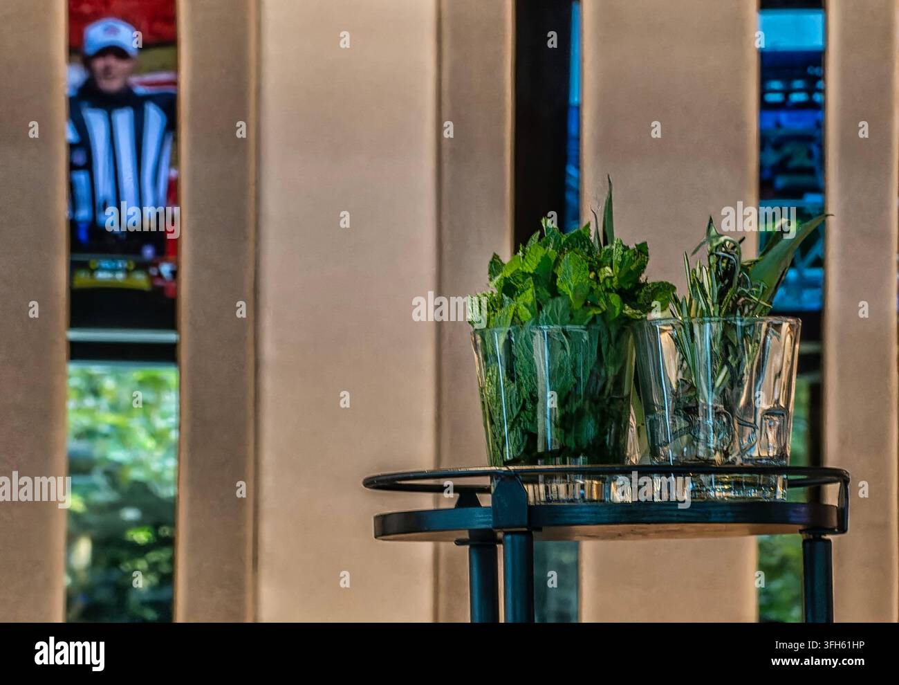 Un elegante carrello bar rotondo e nero in metallo con bicchieri di vetro trasparenti pieni di menta verde fresca ed erbe con riflessi specchiati dallo schermo video del bar Foto Stock