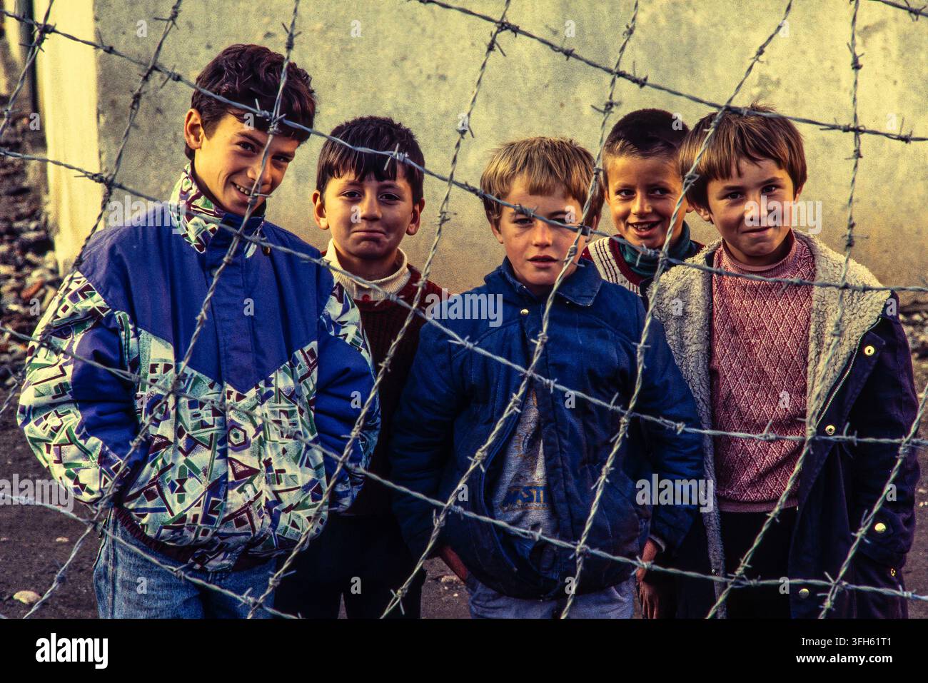 Cinque ragazzi albanesi scrutano attraverso una recinzione di filo spinato, l'infanzia incorniciata dalle barriere di un paese che esce dall'isolamento. Albania, Europa Foto Stock