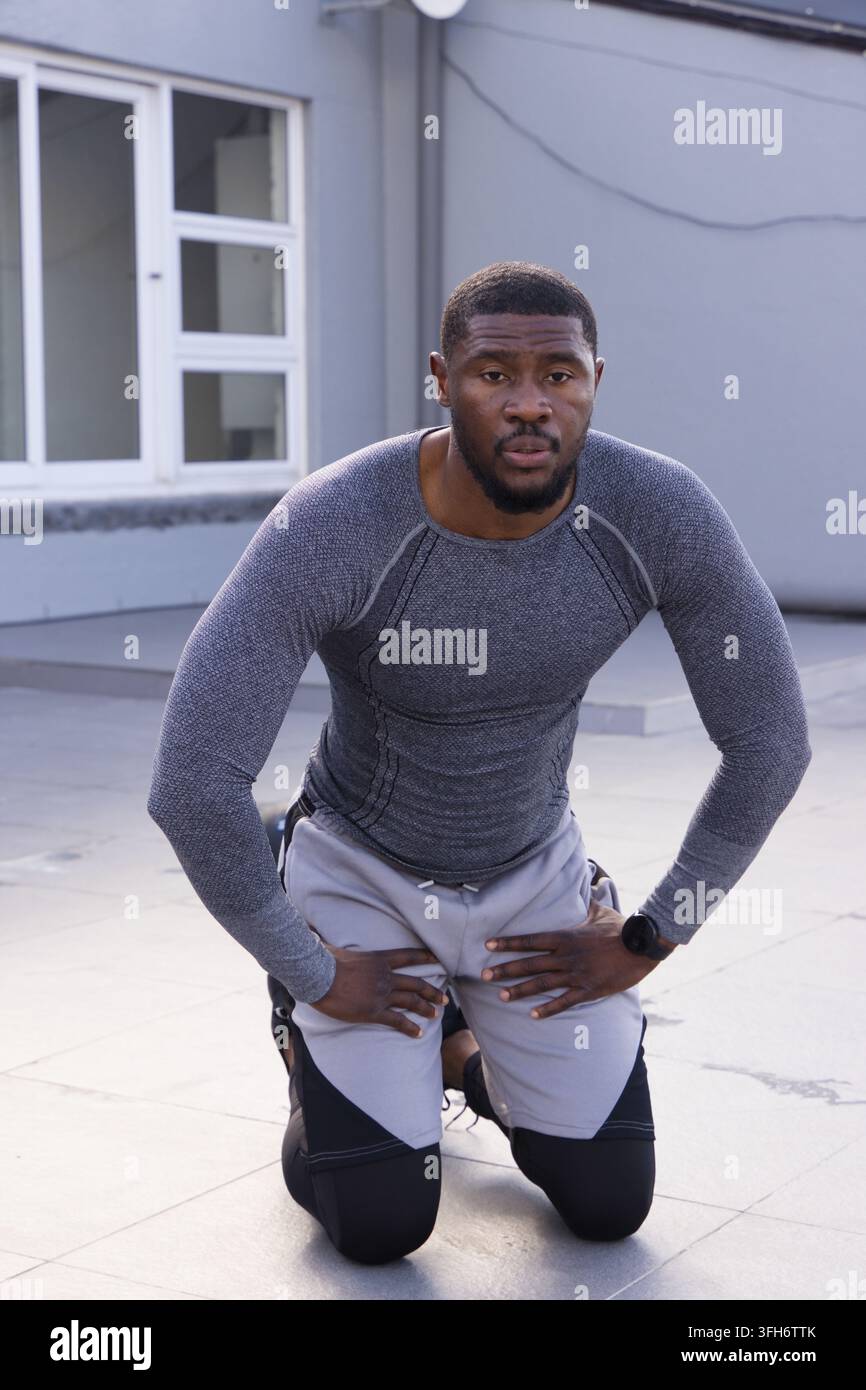 Uomo afroamericano di mezza età inginocchiato sul parapetto del tetto, indossa leggings e controlla lo smartwatch Foto Stock