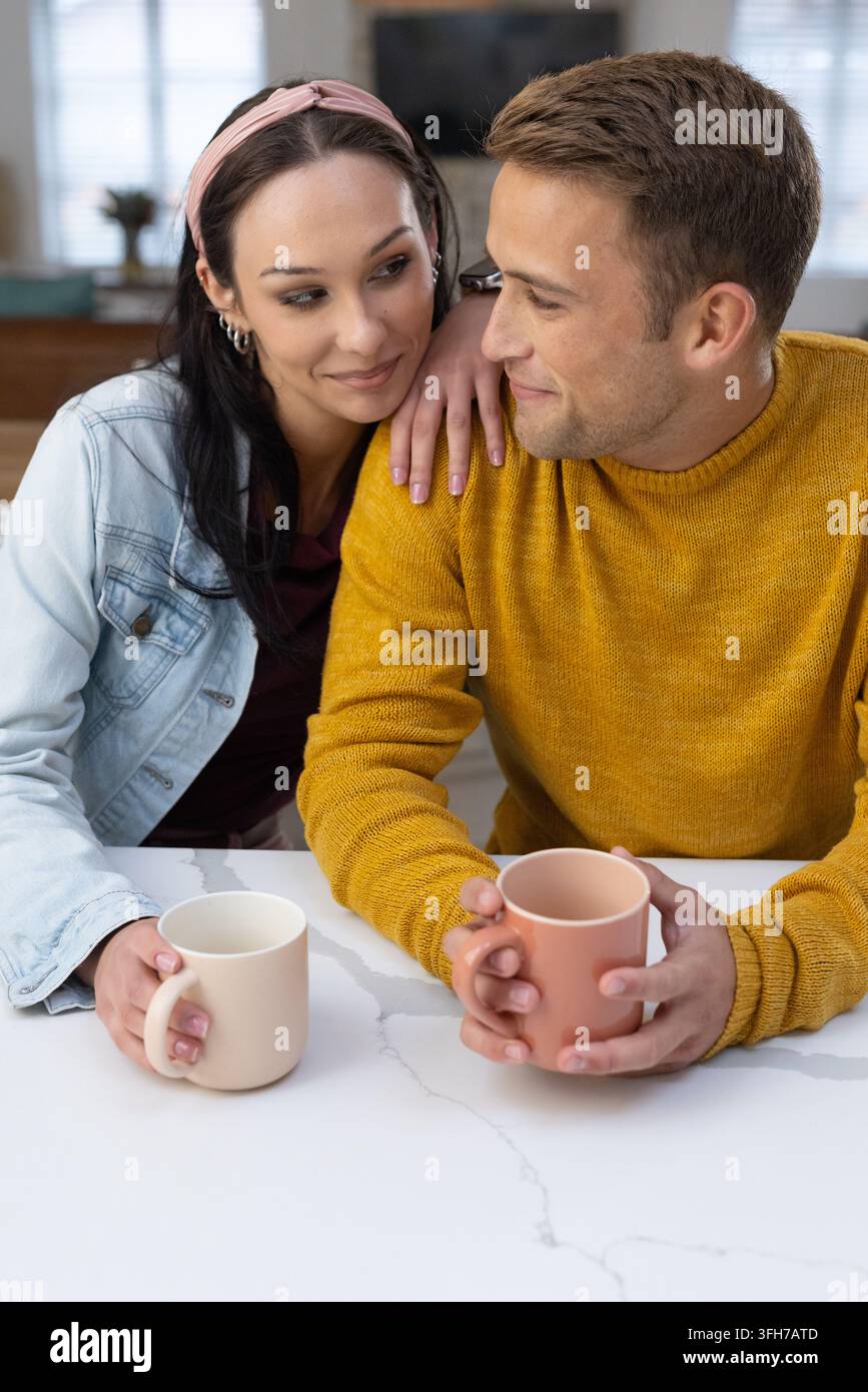 Coppia seduta vicina al bancone di marmo in cucina, indossa maglia senape e giacca di jeans con tazze Foto Stock