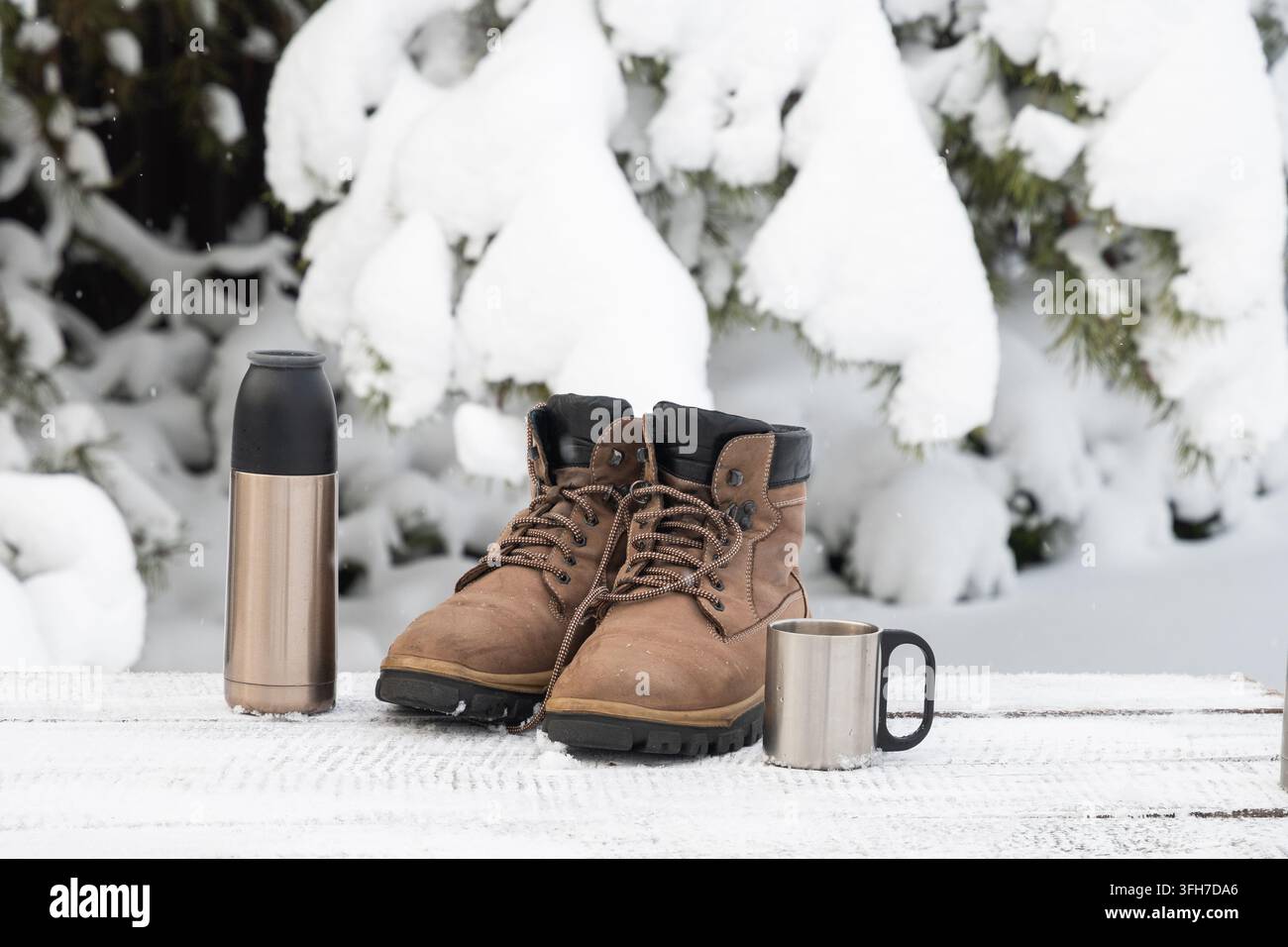 Stivali da trekking, thermos e tazza di metallo con bevanda calda all'aperto in inverno. Concetto di viaggi in condizioni di freddo, pausa riscaldante e svago invernale. Foto Stock