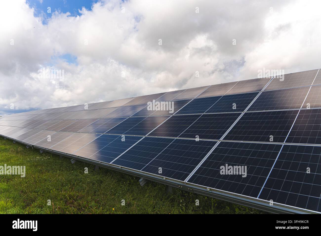 Filiera di pannelli solari fotovoltaici blu che riflettono le nuvole sopra un campo erboso, installazione di energia rinnovabile sotto un cielo parzialmente nuvoloso. Alta qualità p Foto Stock