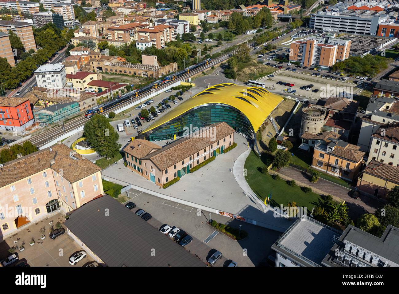Vista aerea del Museo Enzo Ferrari con il suo tetto giallo iconico e la moderna facciata in vetro adiacente ad edifici storici e binari ferroviari a Modena, Foto Stock