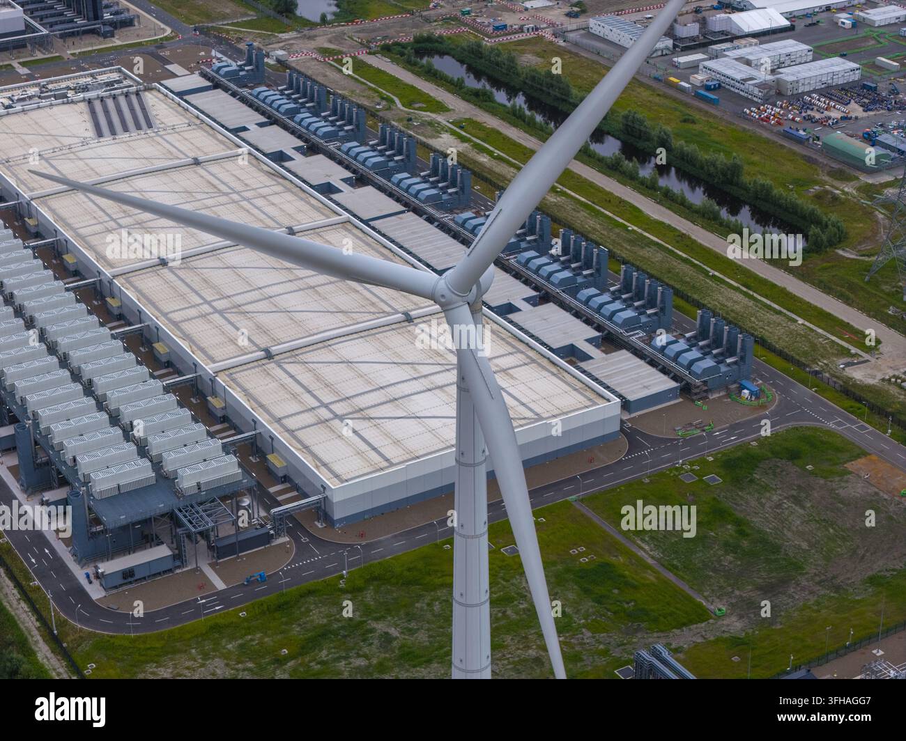 Vista aerea di un grande centro dati con una prominente turbina eolica in primo piano, circondata da infrastrutture industriali e paesaggio verde Foto Stock