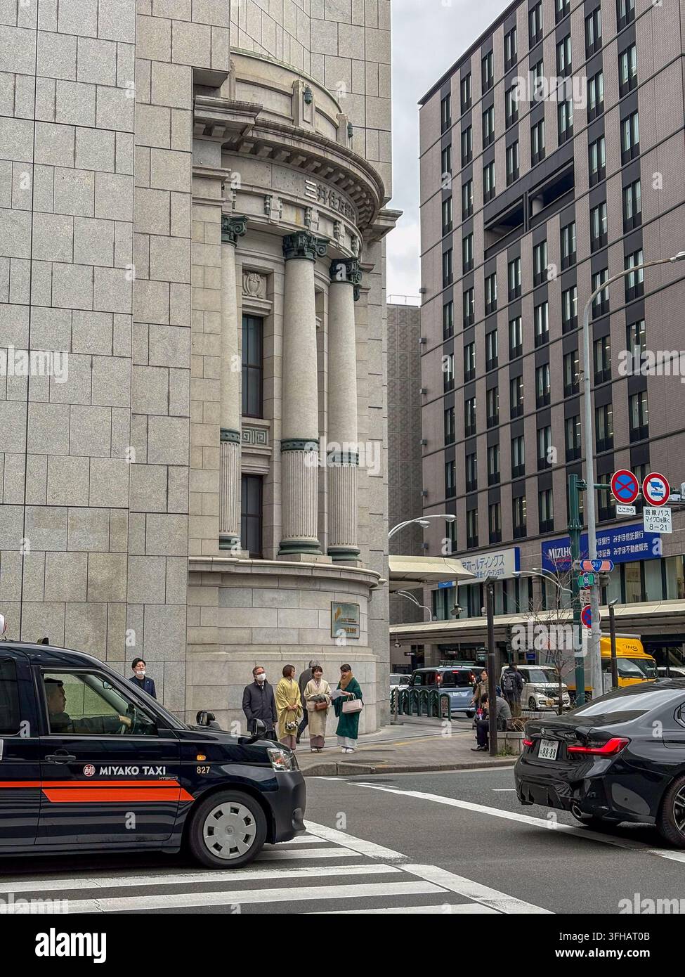 Classiche colonne in pietra su una facciata di edificio storico con persone che indossano kimoni tradizionali su un attraversamento pedonale a Kyoto. - Immagine stock catturata con smartphone