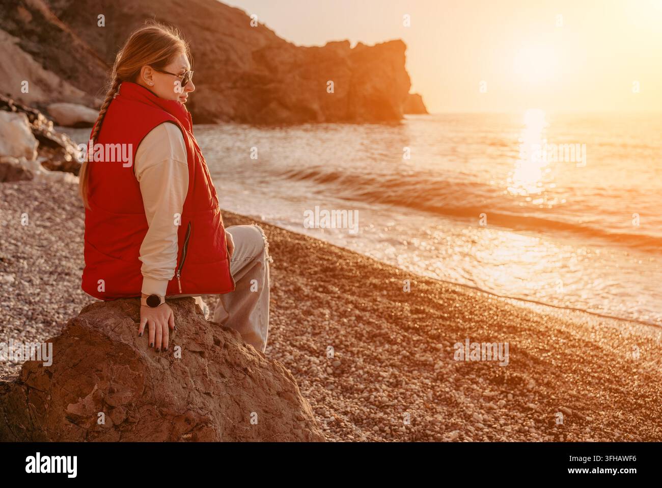 Donna seduta su una roccia sulla spiaggia al tramonto che osserva il sole dorato immergersi sotto l'orizzonte durante una serata tranquilla in riva al mare Foto Stock
