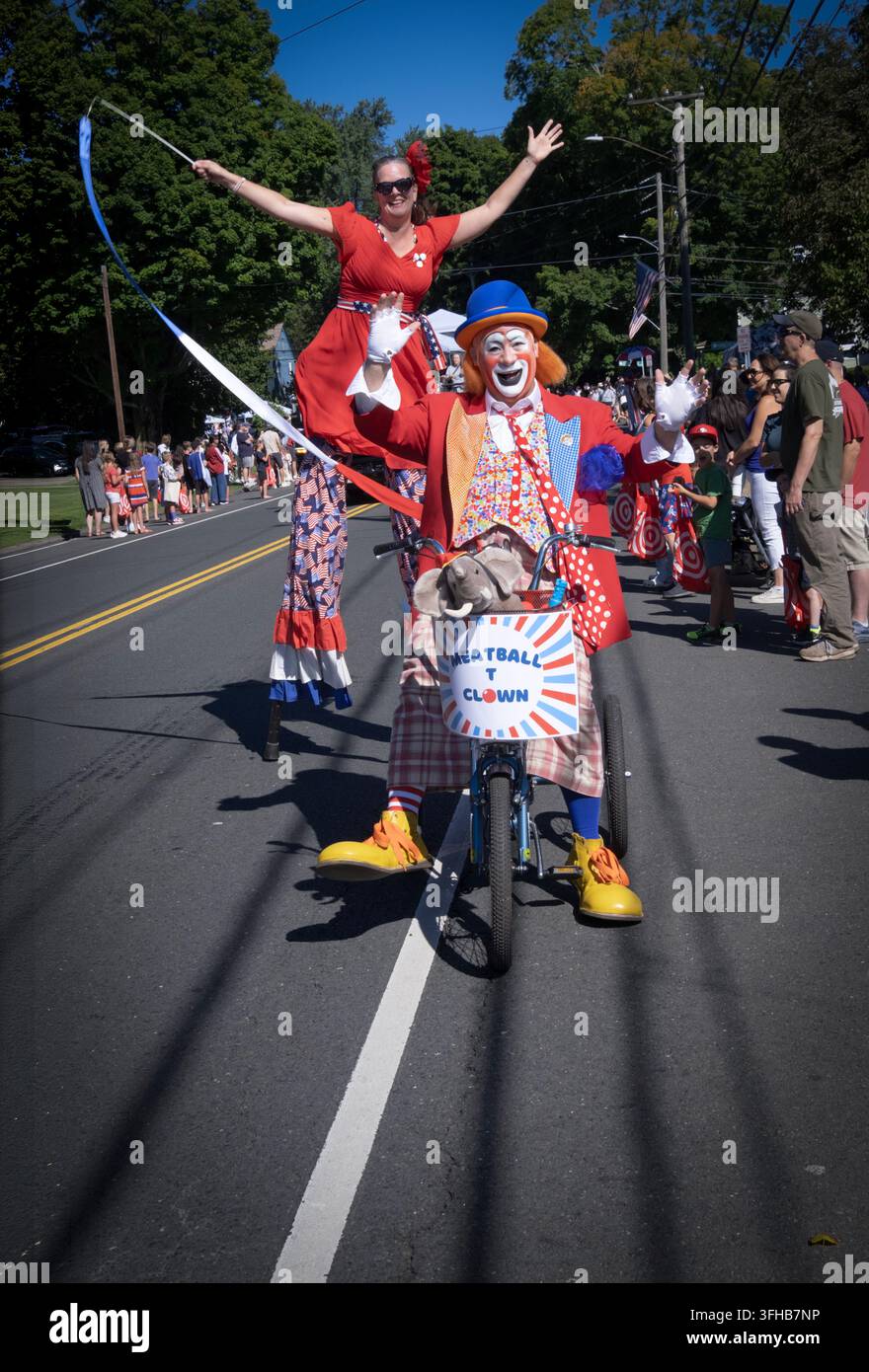 Con un camminatore su palchi sullo sfondo, il clown Meatball T pedala su Main Street alla parata del Labor Day di Newtown nel 2026 nel Connecticut. Foto Stock