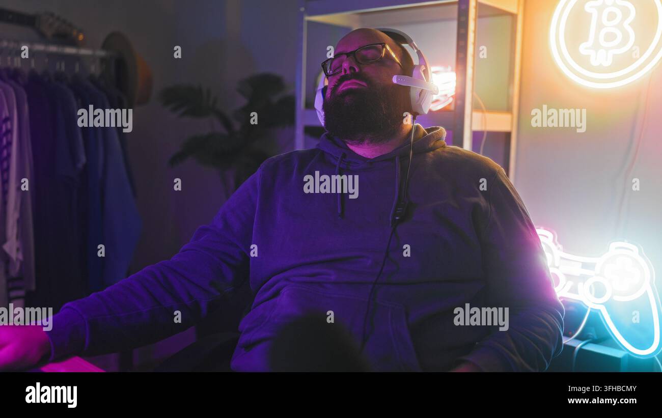 Uomo con barba e occhiali che indossa un auricolare si appoggia con gli occhi chiusi alla scrivania del computer accanto a un cartello al neon del controller e uno scaffale per vestiti nello studio; stanco Foto Stock