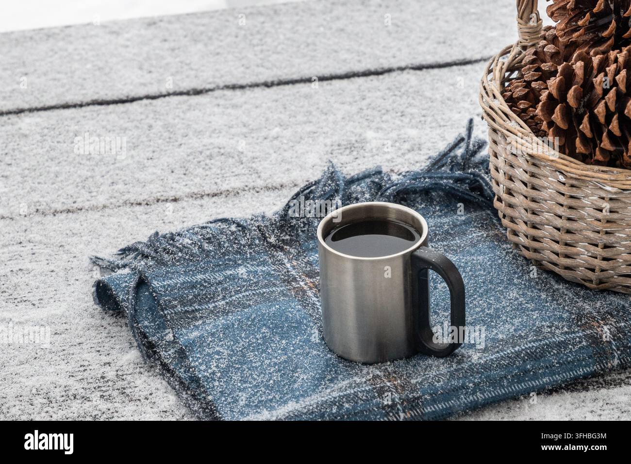 Tazza da campo in metallo piena di bevanda calda si trova su una coperta a quadri blu coperta di neve fresca accanto a un cesto di pigne, con spazio per il testo. Foto Stock