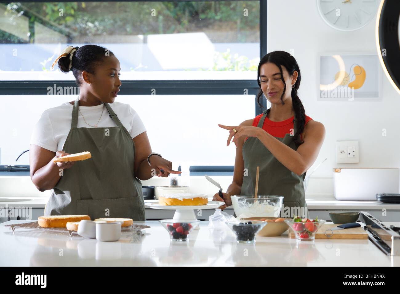 Diverse amiche con grembiuli verdi che assemblano una torta a più piani in una cucina moderna con le spatole Foto Stock
