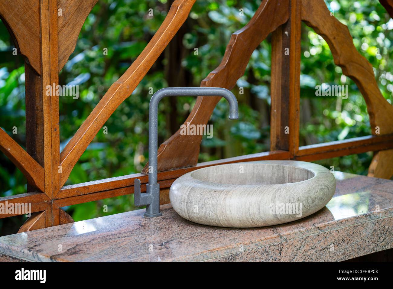 Lavandino moderno in pietra e rubinetto in interno esterno in legno. Elegante lavabo rotondo in pietra e rubinetto grigio opaco su piano di lavoro in granito davanti al legno Foto Stock