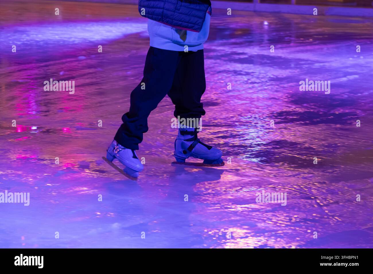Piedi in pattini da ghiaccio blu scivolano su una pista riflettente, illuminata da vivaci luci al neon viola e rosa, catturando il tempo libero invernale, lo spirito delle festività e Foto Stock