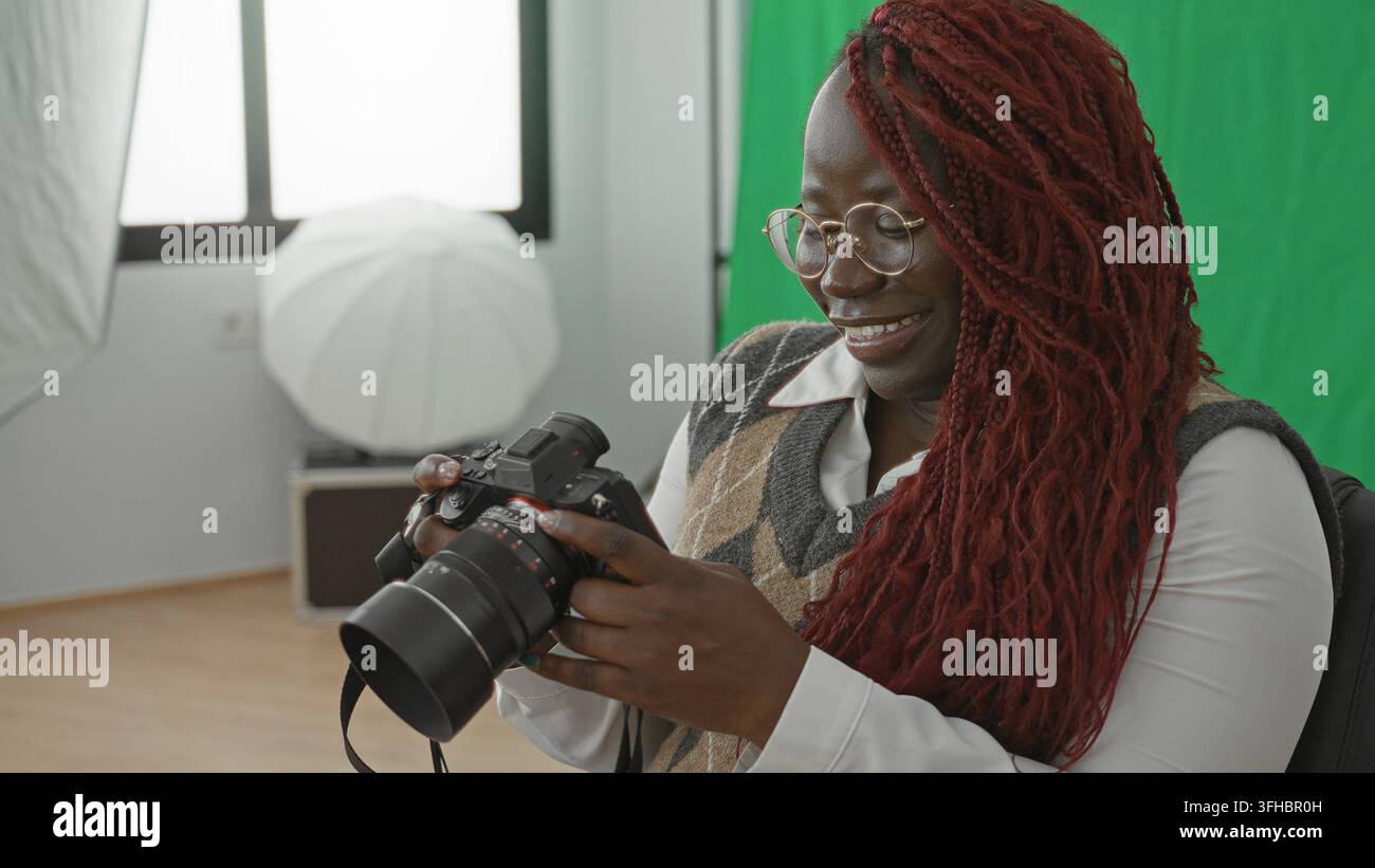 Donna tiene la fotocamera e controlla le impostazioni in studio con schermo verde, softbox e finestra visibili; concentrazione creatività. Foto Stock