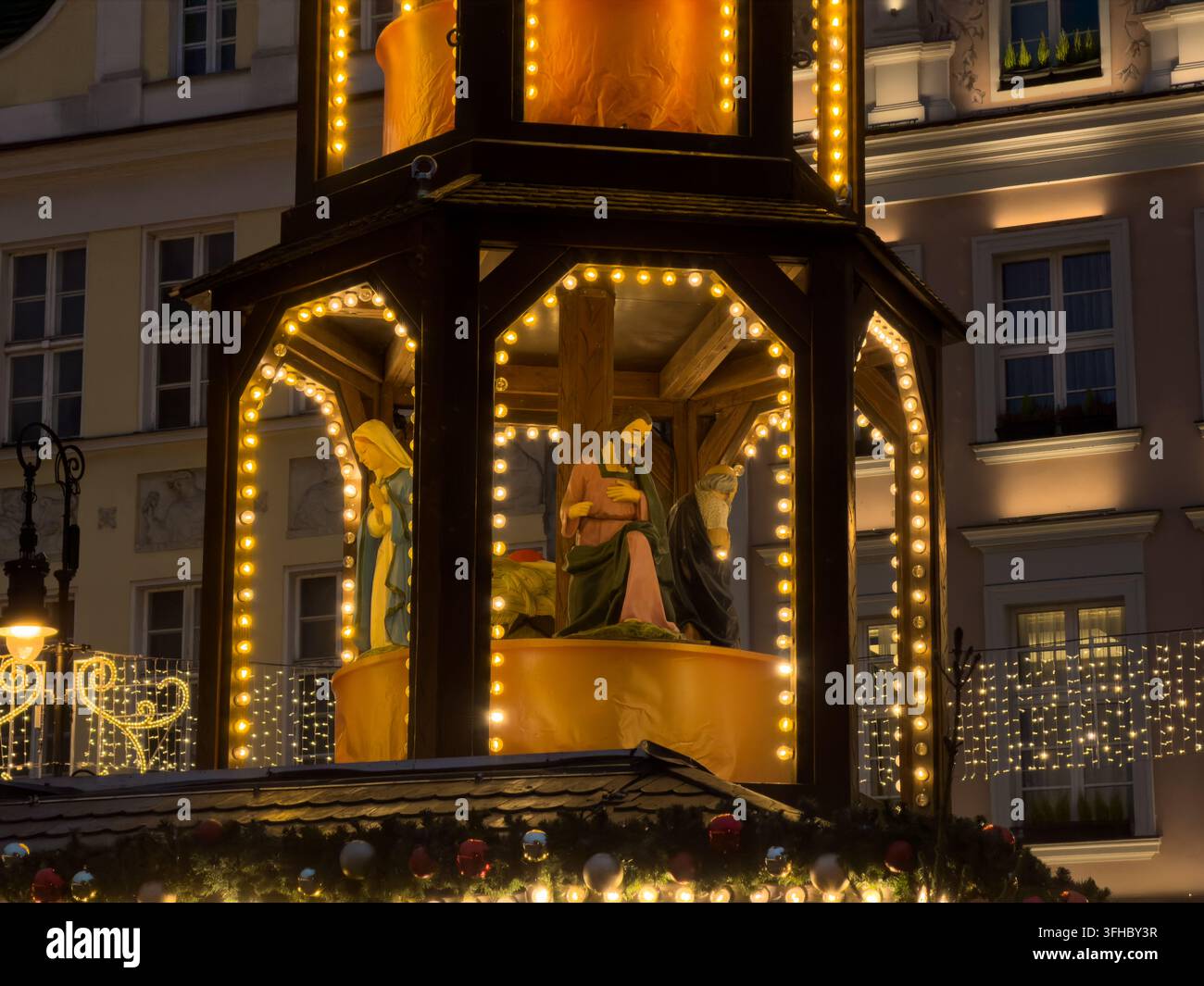 Pozna, Polonia – 13 dicembre 2025: La scena della natività in legno mostra figure luminose sotto luci dorate in un mercato all'aperto, celebrando la stagione Foto Stock