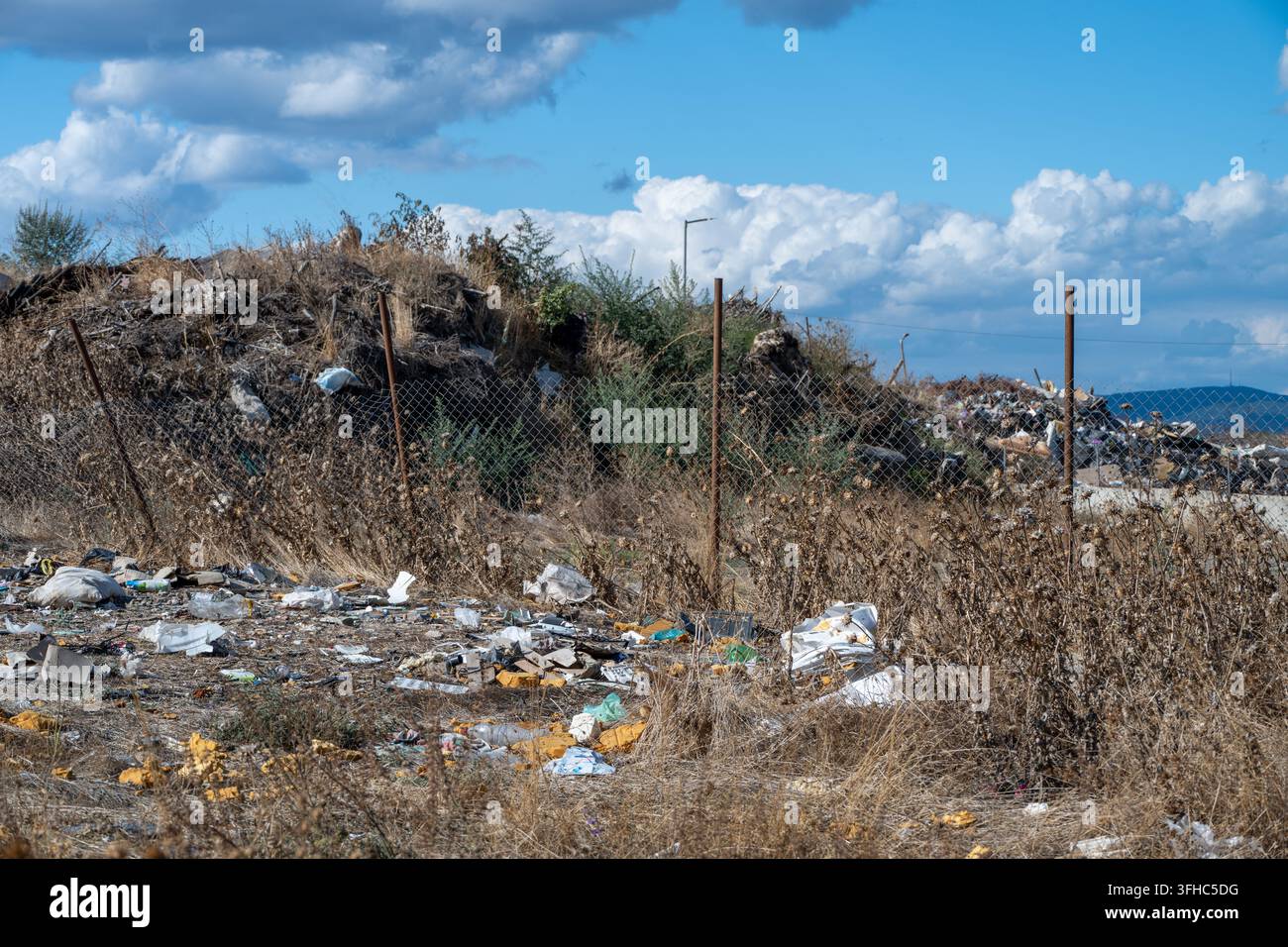Rifiuti di plastica sparsi e immondizia ricoprono il terreno secco e invaso dalle erbacce fuori da una recinzione di filo metallico che delimita una discarica aperta e cumuli di compost di rifiuti organici b Foto Stock