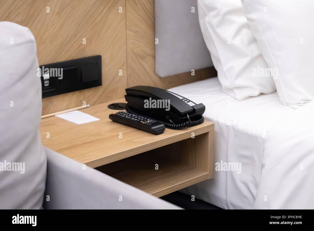 Un auricolare nero del telefono riposa accanto a un telecomando su un comodino minimalista in legno, offrendo comodità agli ospiti in un ambiente ordinato e ben progettato Foto Stock