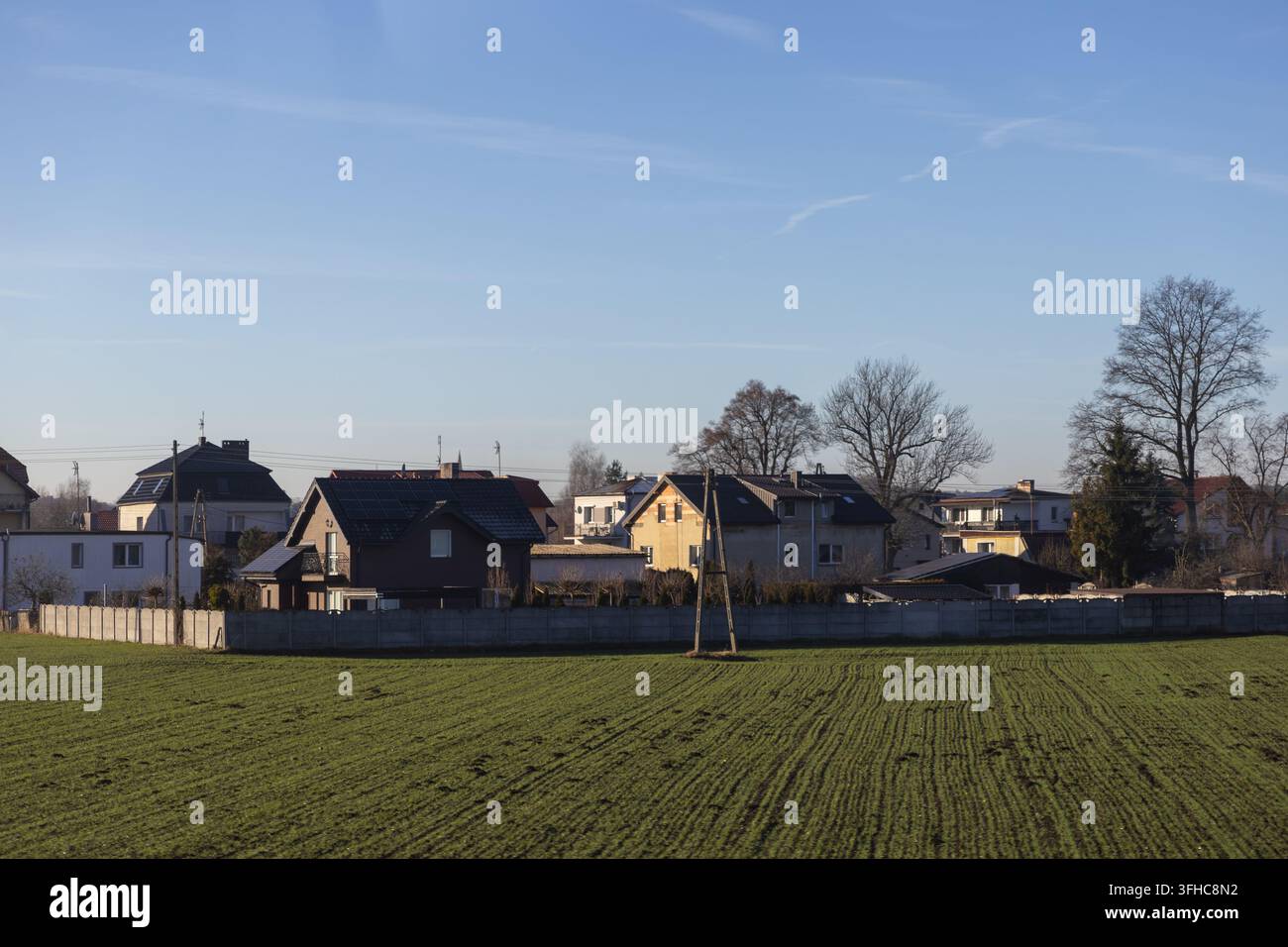 Edifici residenziali silenziosi si trovano vicino a un terreno agricolo aperto separato da un'alta barriera, illustrando una miscela di stile di vita rurale e natura pastorale Foto Stock
