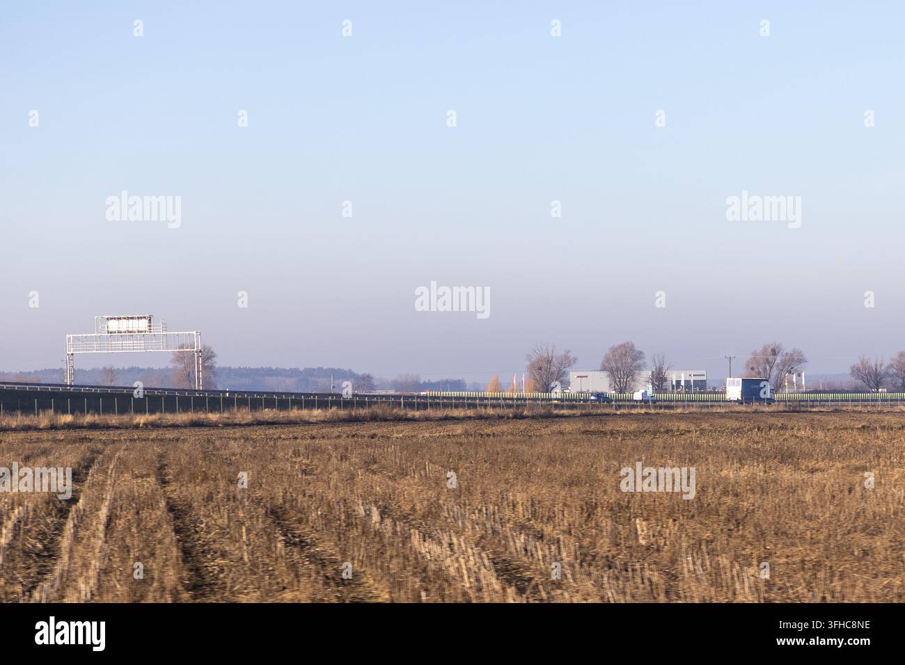 Le spighe dorate di un campo agricolo arato si estendono davanti a una moderna autostrada, catturando il netto contrasto tra il ciclo tradizionale della raccolta stagionale Foto Stock