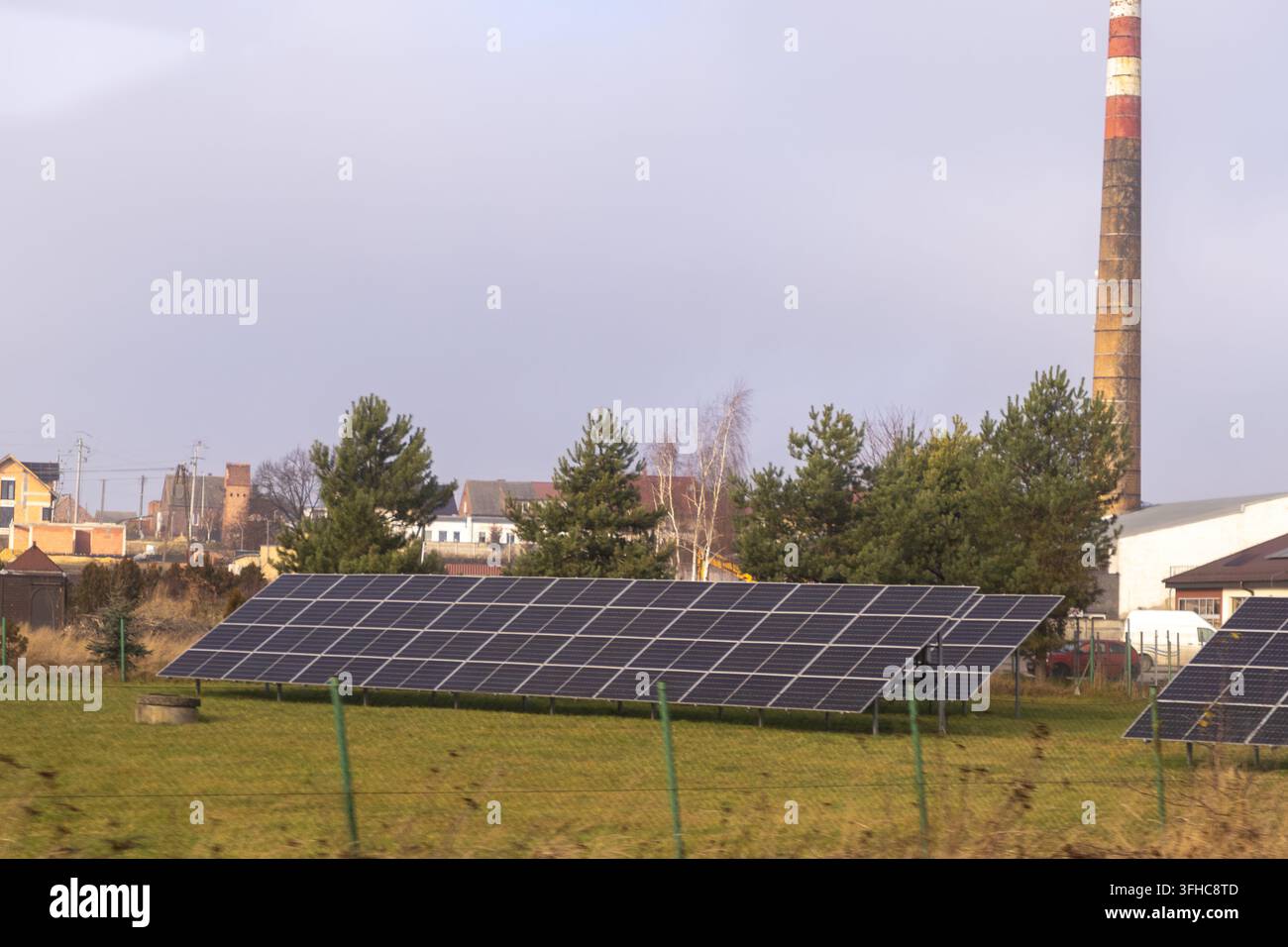 File di pannelli fotovoltaici si ergono in un campo aperto sullo sfondo di alberi e un camino industriale, illustrando la transizione verso l'energia sostenibile Foto Stock