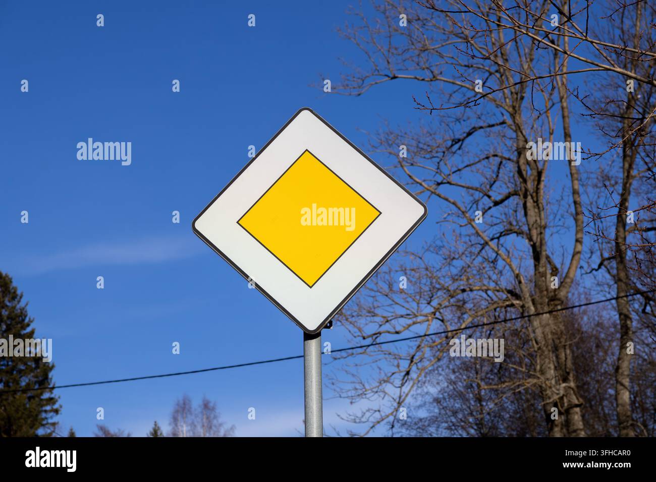 Una vista dettagliata di un cartello di priorità della strada principale, con un rombo bianco e un quadrato giallo su un cielo blu vivace, simbolico delle regole critiche del traffico Foto Stock