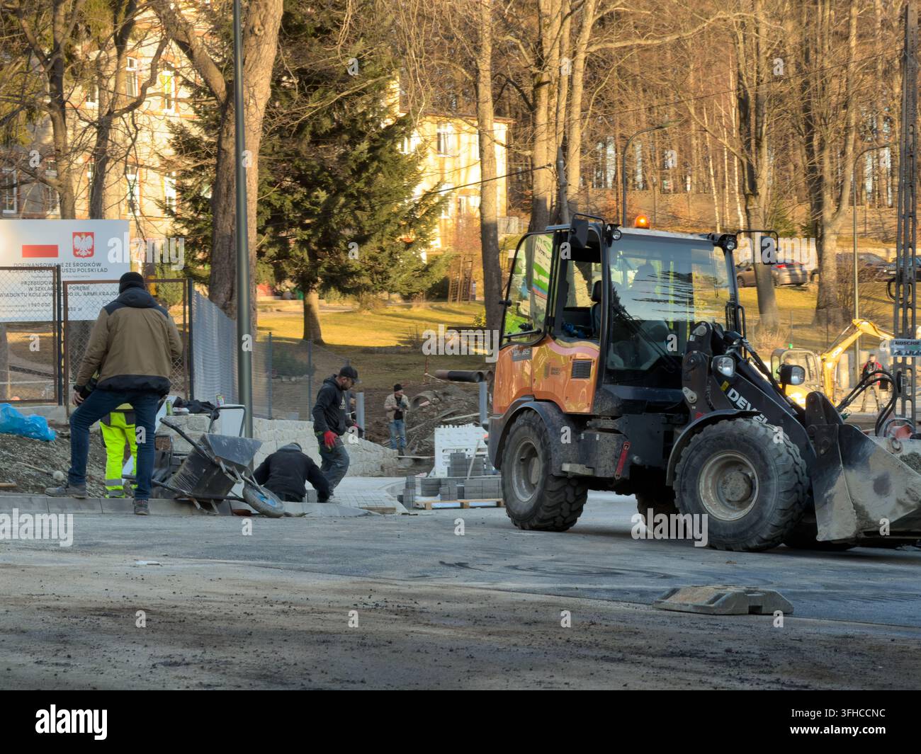 Szklarska Poręba, Polonia – 16 dicembre 2025: Diversi operai edili posano diligentemente pietre di pavimentazione per un nuovo progetto stradale urbano, supportati da un c Foto Stock