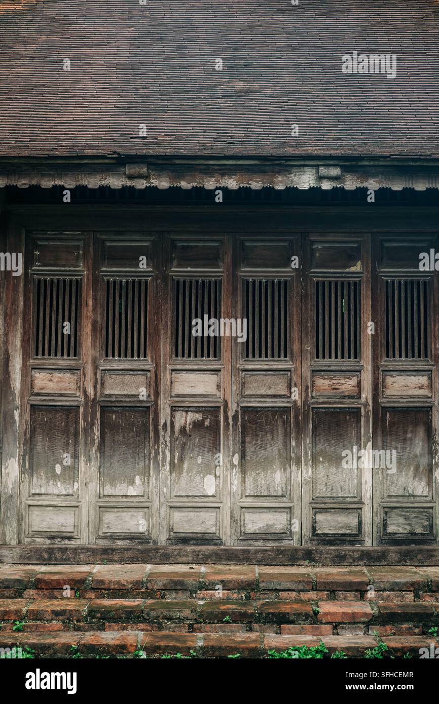 Hue, Vietnam - 9 settembre 2026. Porte di legno consumate riempiono la facciata di un edificio storico sotto un tetto di tegole invecchiato e gradini di mattoni. Foto Stock