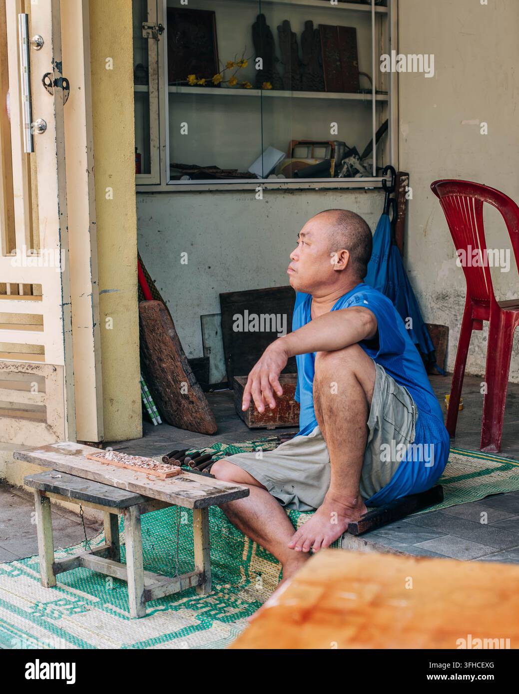 Hue, Vietnam – 9 settembre 2026. Un uomo di mezza età è seduto scalzo fuori da una casa modesta accanto a un piccolo tavolo, un tappeto, una sedia e oggetti domestici. Foto Stock