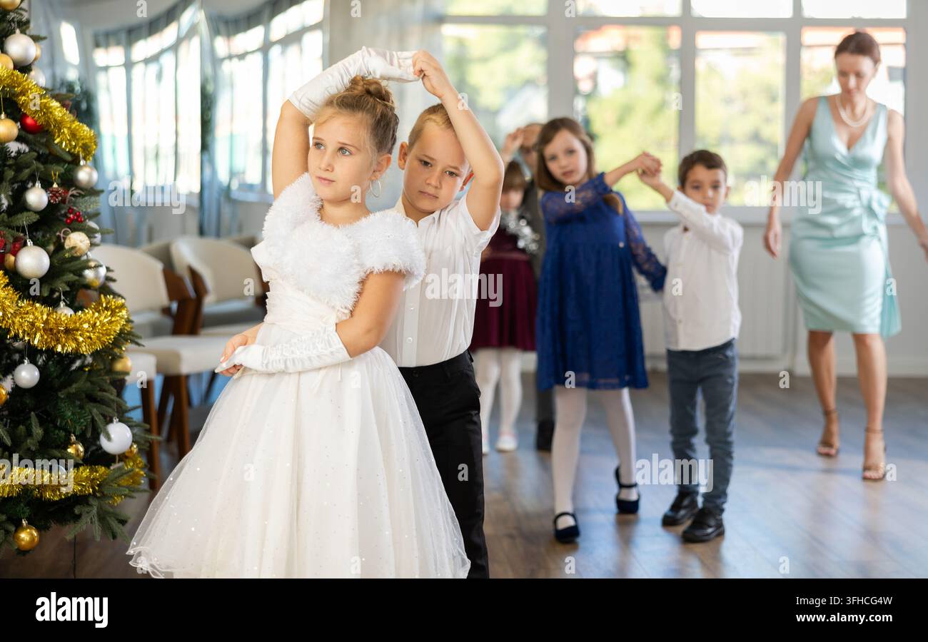 Preadolescenti entusiasti, ragazze e ragazzi in abiti eleganti che praticano passi di danza di coppia in uno studio adornato di decorazioni natalizie, sotto Gu Foto Stock