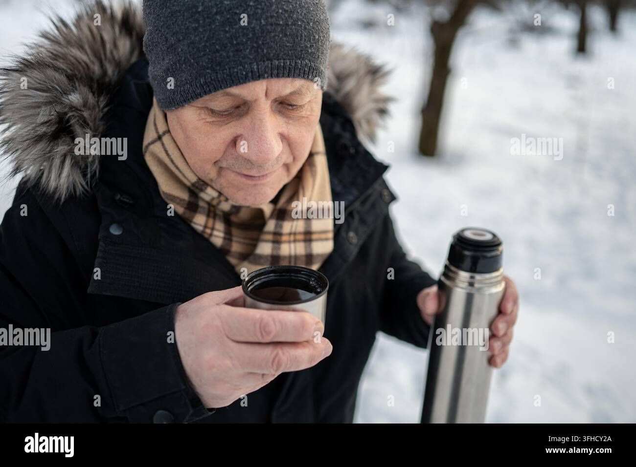 Uomo caucasico maturo che annusa o si appresta a bere una bevanda calda da una tazza termica all'aperto durante una passeggiata invernale nella neve fredda. Foto Stock