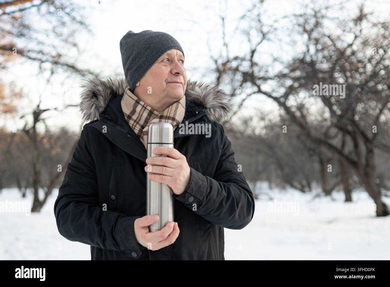 Uomo caucasico maturo che tiene un thermos di metallo durante una passeggiata in un parco innevato, preparato con una bevanda calda per il freddo inverno. Foto Stock