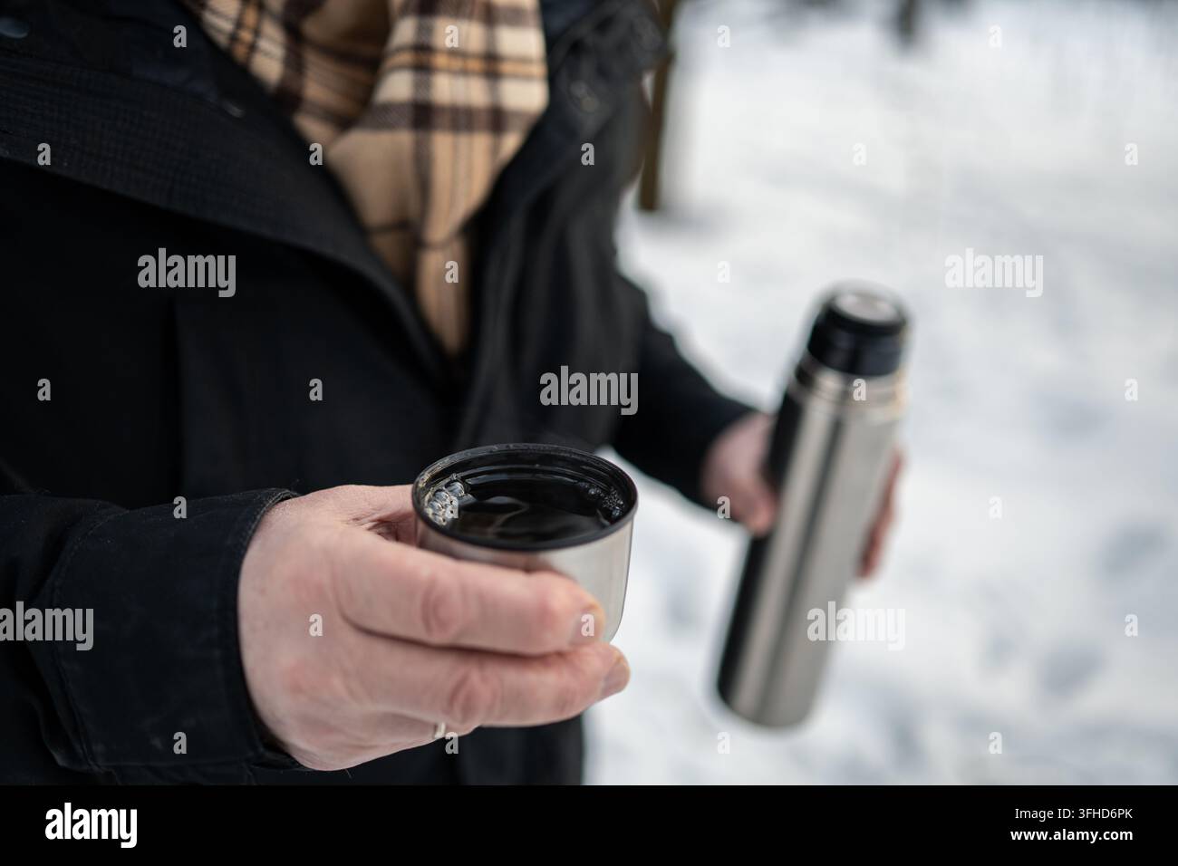 Primo piano di un uomo maturo che tiene una tazza di bevanda calda e una thermos in metallo all'aperto nella neve, riscaldandosi con una bevanda durante il freddo inverno. Foto Stock