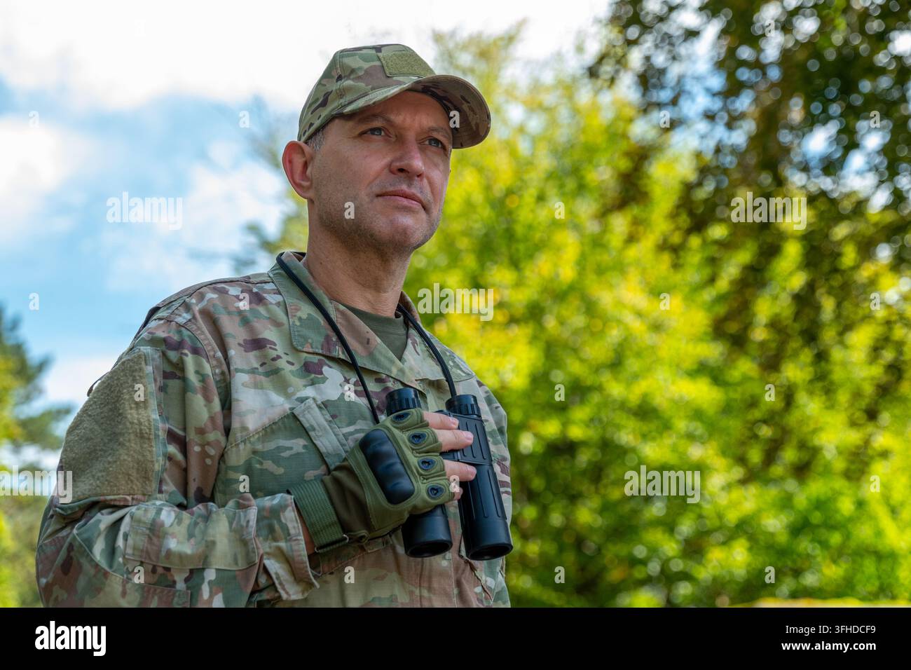 Un uomo in abbigliamento mimetico tiene un binocolo mentre è all'aperto contro uno sfondo sfocato di foresta verde. Foto Stock