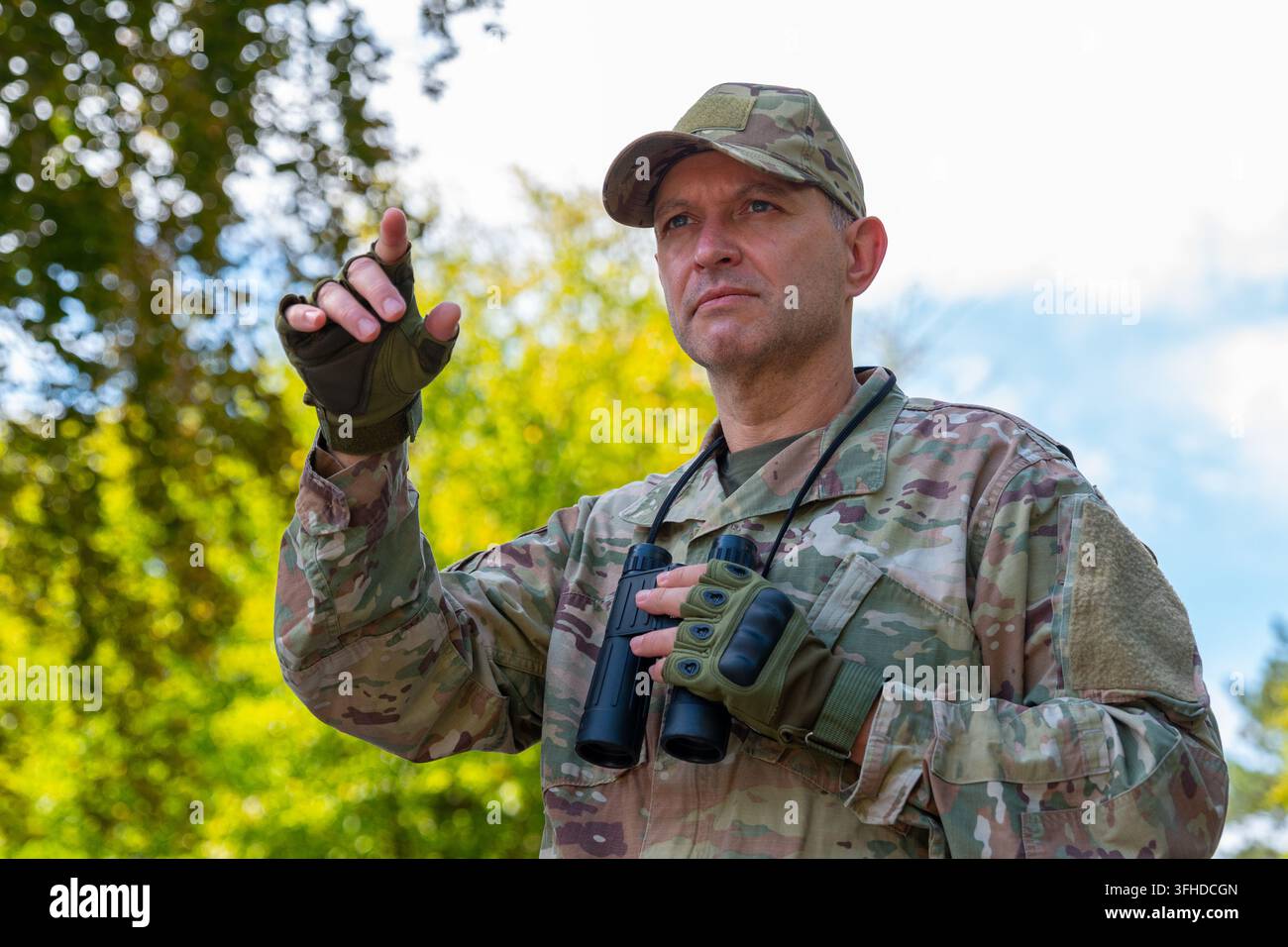 Un uomo in uniforme mimetica indica verso l'alto mentre tiene il binocolo in un ambiente esterno luminoso. Foto Stock