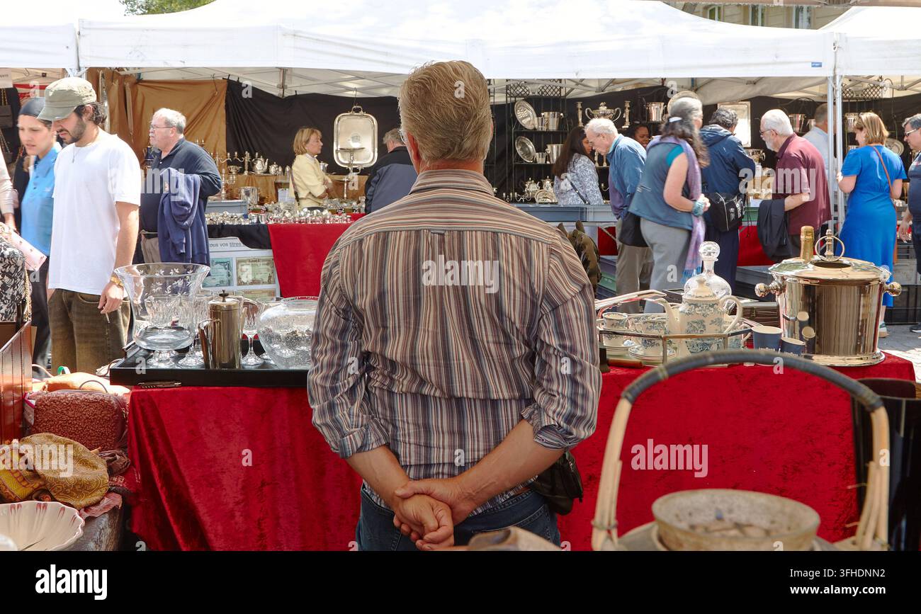 Parigi, Francia – 08 maggio 2026: una vista posteriore di un uomo che osserva stoviglie vintage e oggetti da collezione in un affollato mercatino delle pulci di strada Foto Stock