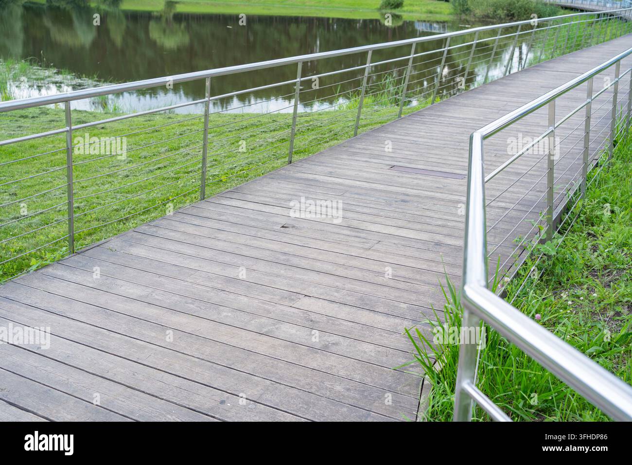 Un tranquillo passaggio di legno in un contesto di parco, che conduce a uno stagno sereno circondato da una vegetazione lussureggiante e toni di terra bruni. La prospettiva in prima persona Foto Stock