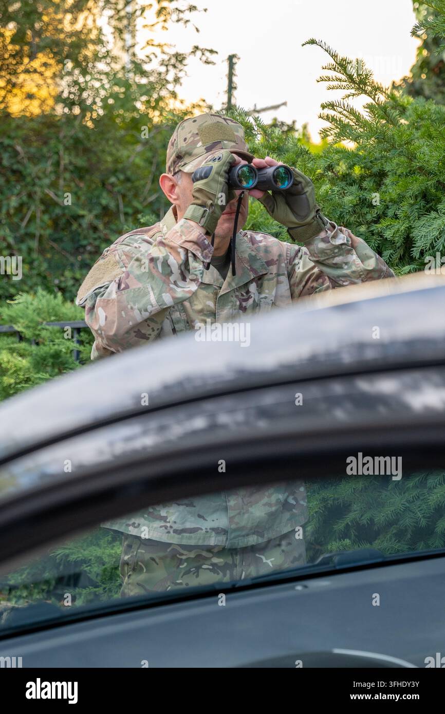 Operativo dell'intelligence militare in mimetica osserva attraverso un binocolo da una posizione nascosta vicino a un veicolo durante un'operazione segreta Foto Stock