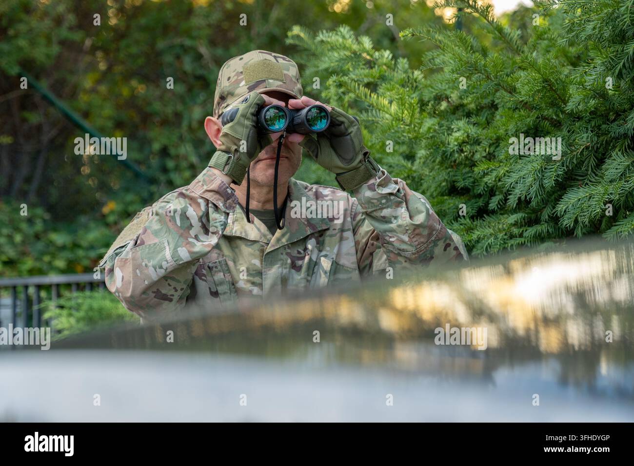 L'agente dell'intelligence monitora un obiettivo distante con un mirino rimanendo nascosto vicino a un veicolo e a vegetazione fitta Foto Stock