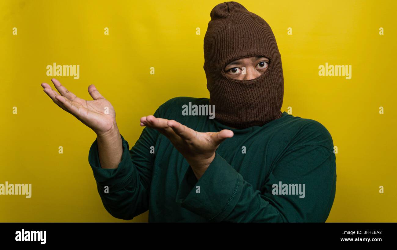 Giovane uomo asiatico con passamontagna marrone alza entrambe le mani in gesto interrogativo su sfondo giallo brillante. L'individuo mascherato mostra conf Foto Stock