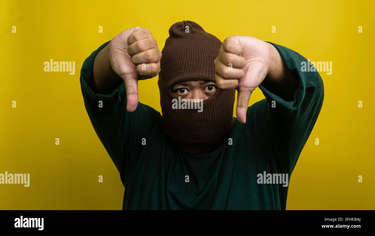 Giovane uomo asiatico con passamontagna marrone mostra gesto a due pollici verso il basso su uno sfondo giallo vivace. L'individuo mascherato proietta una forte disapprovazione Foto Stock